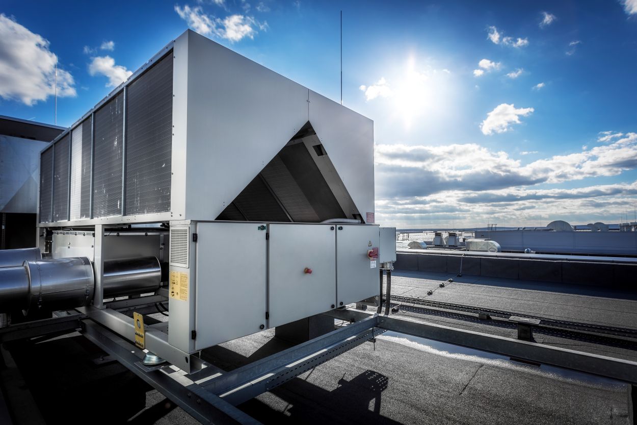 A huge air conditioning unit on the roof of the building. In the background of blue sky with shining sun. Focus is at the front of the air conditioner, the other parts of image slightly blurred.